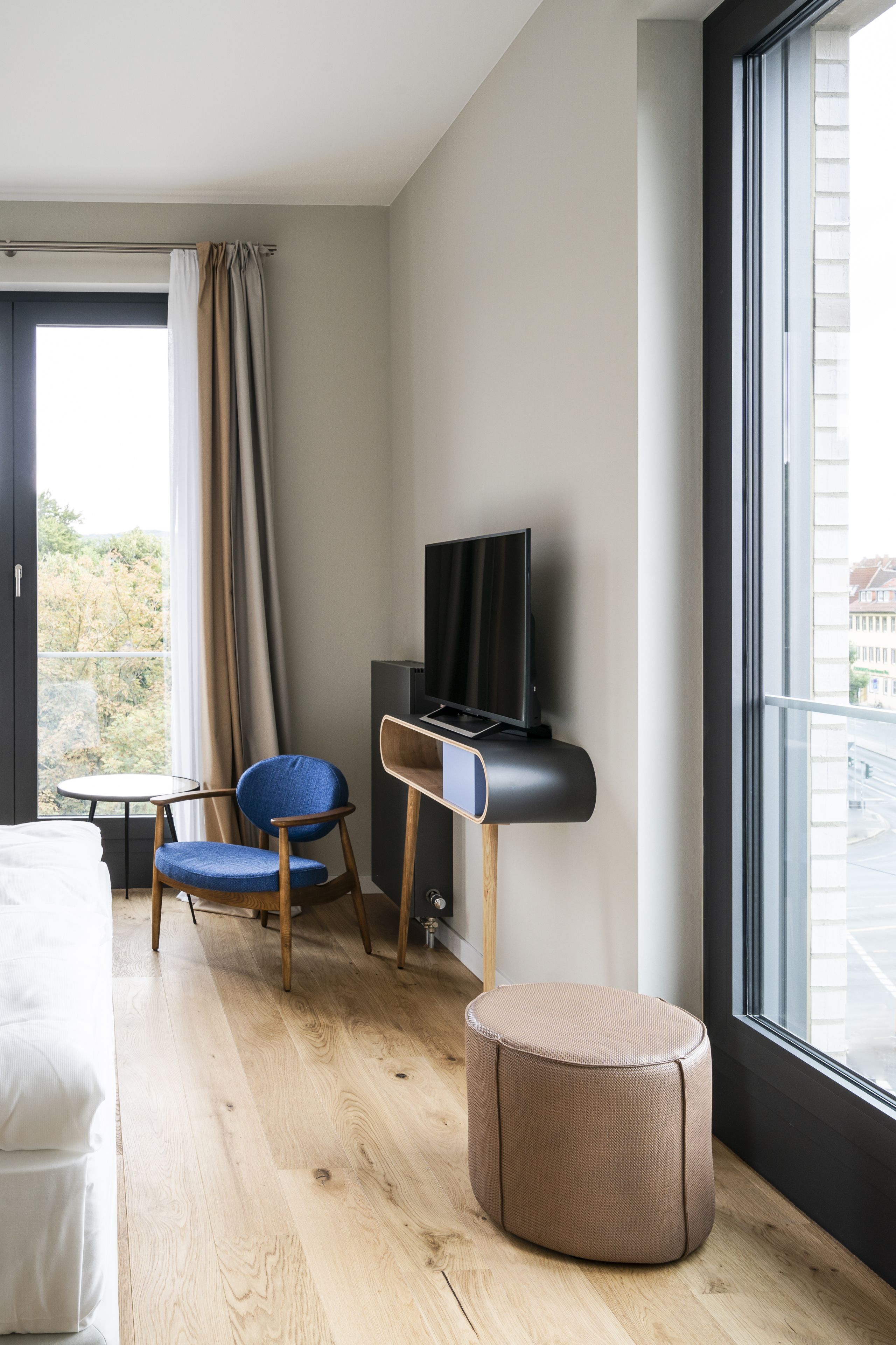 Habitación de hotel moderna con cama, televisor sobre un soporte, una silla, una mesita y un amplio ventanal con vistas a edificios.