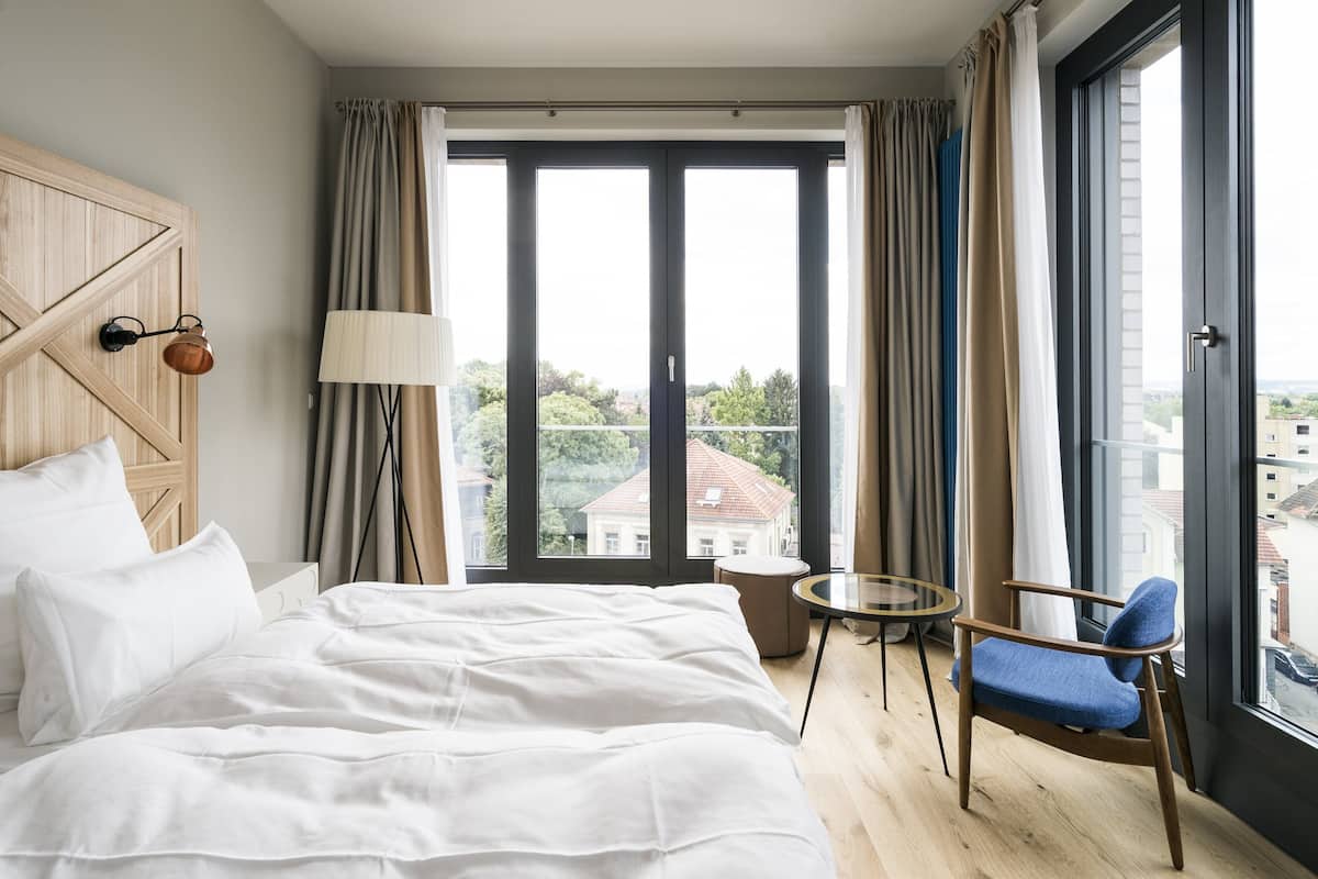 A hotel room with a large bed, a small round table, a chair, and a view of buildings through a large window.