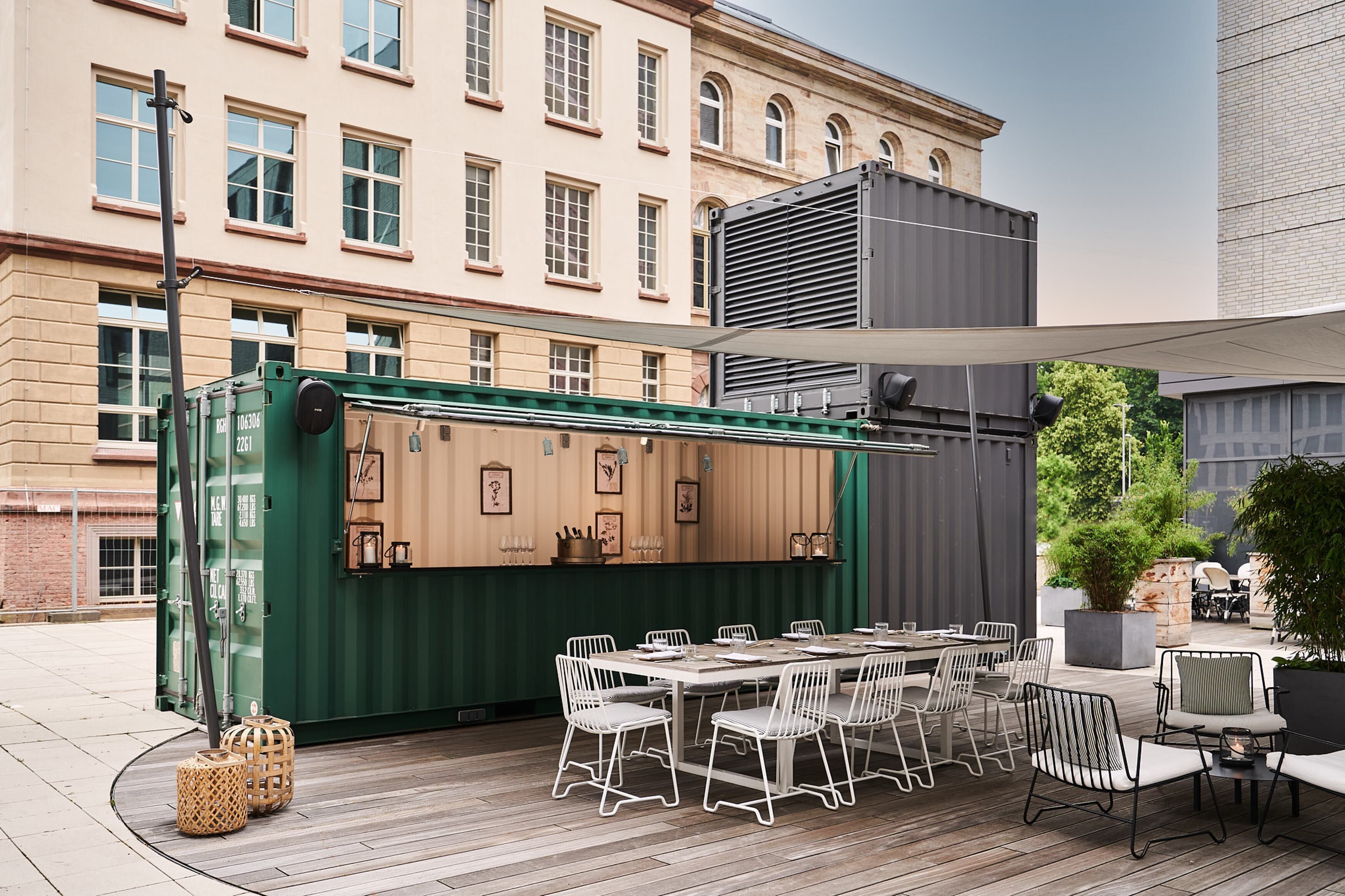 Un área de comedor al aire libre con una mesa larga y sillas, un contenedor verde con equipamiento de cocina y una vista de un edificio histórico al fondo.