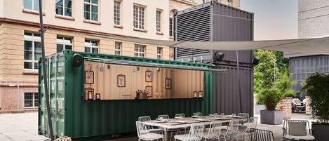Un área de comedor al aire libre con una mesa larga y sillas, un contenedor verde con equipamiento de cocina y una vista de un edificio histórico al fondo.