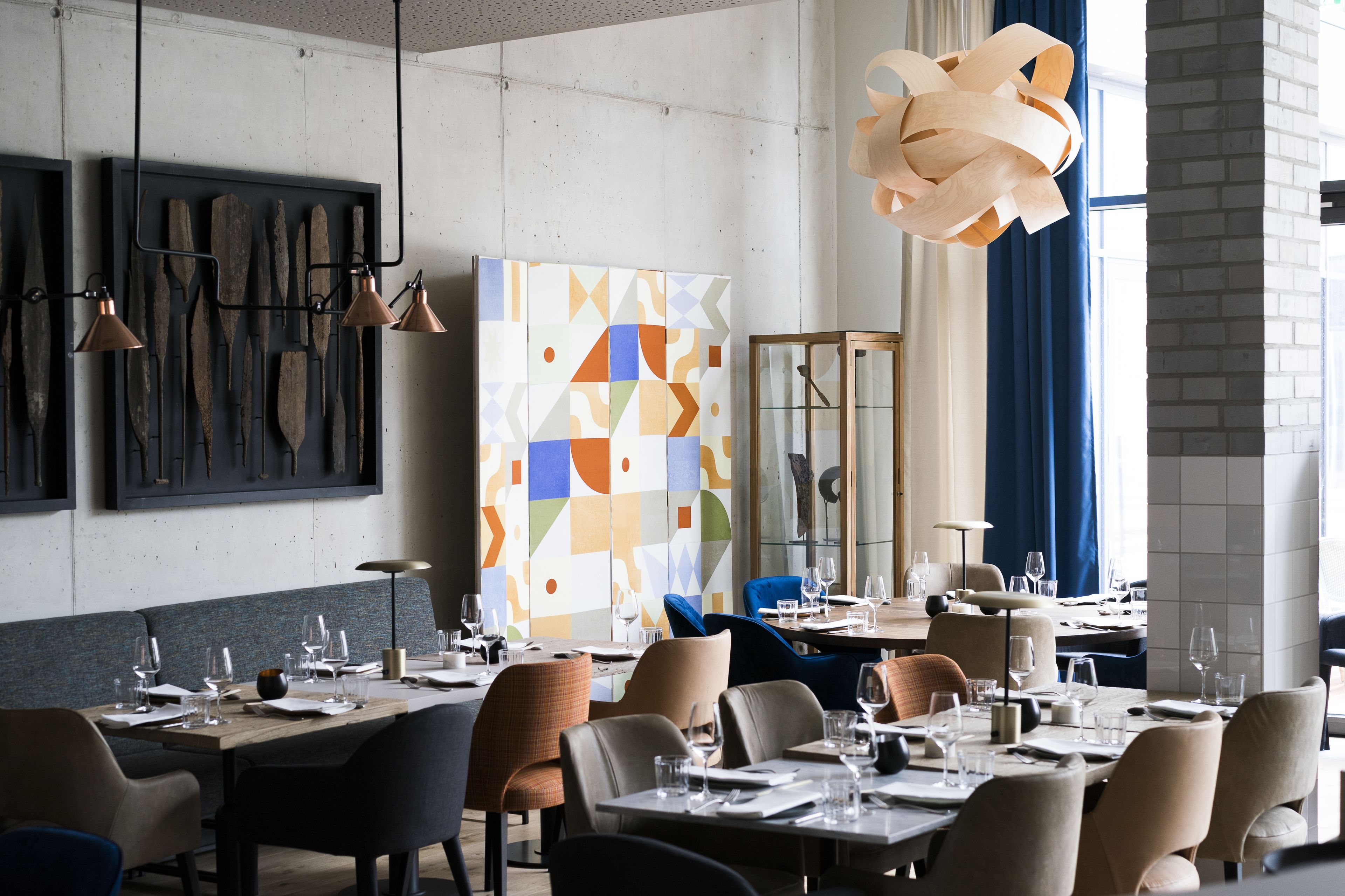 Un interior de restaurante moderno con mesas dispuestas para comer, una obra de arte geométrica en la pared y una gran lámpara de madera.