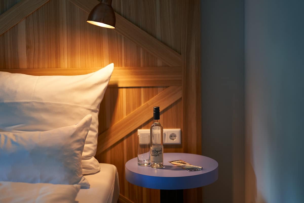 A bed with white pillows, a bottle of alcohol, and a glass on a small round table with a blue surface.