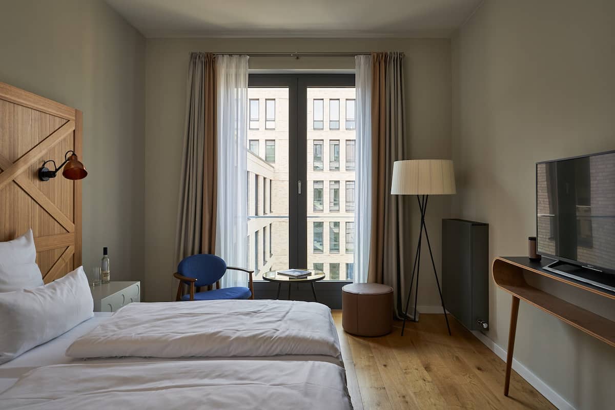 A hotel room with a bed, a chair, a small table, a lamp, a TV, and a window with curtains.