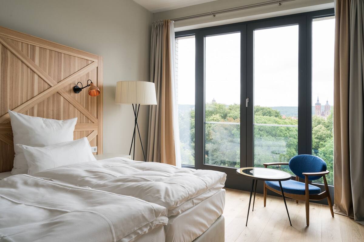 A hotel room with a large bed, a chair, a lamp, and a view of greenery and buildings through the window.
