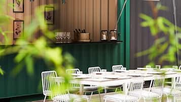 A dining area with white metal chairs and tables, a green awning, and a building with a window and a door in the background.