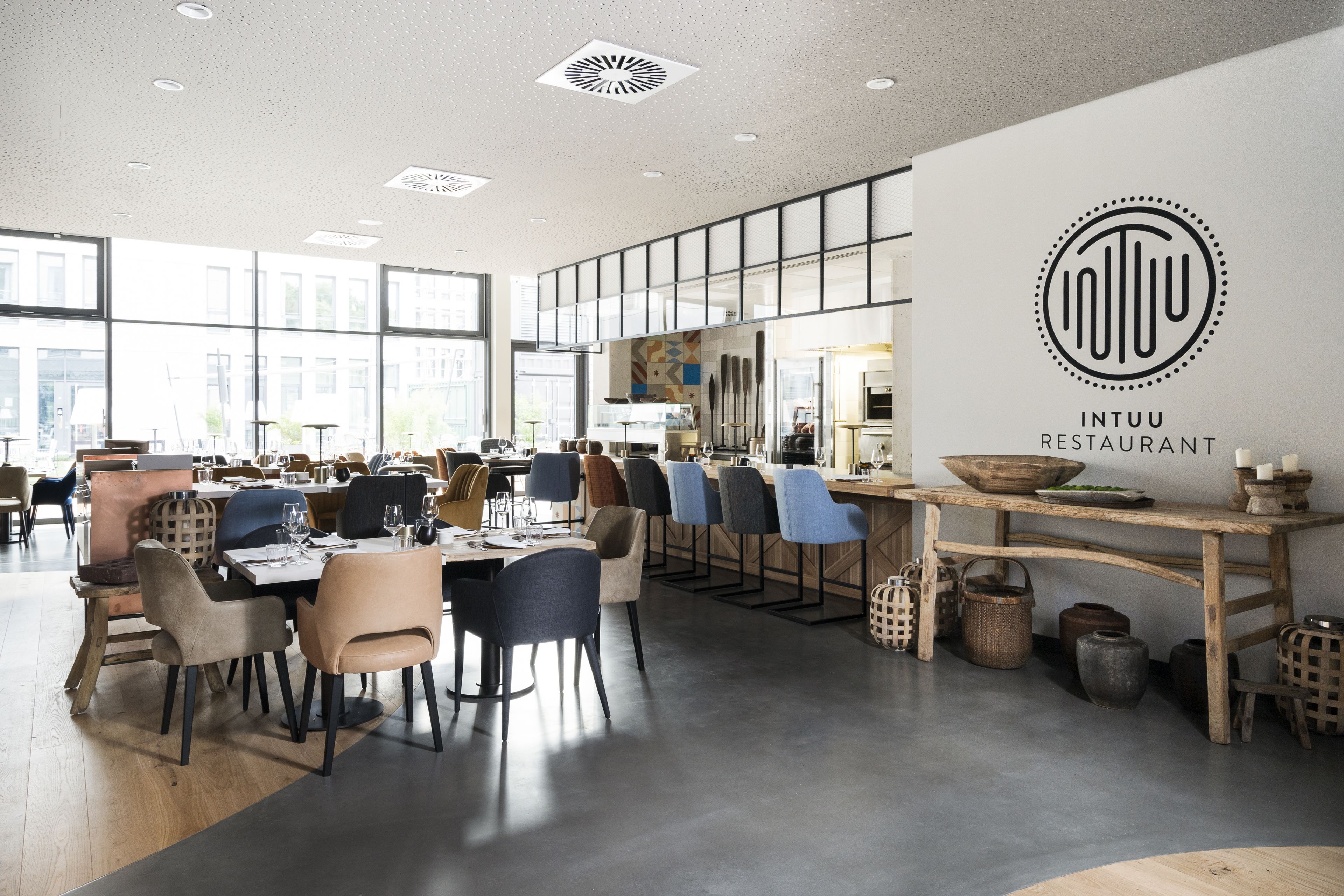 Et moderne restaurantinteriør med høje lofter, store vinduer og et centralt spiseområde med flere borde og stole.