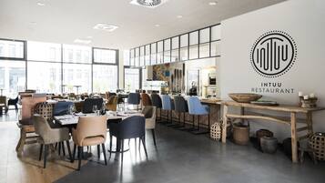 Et moderne restaurantinteriør med høje lofter, store vinduer og et centralt spiseområde med flere borde og stole.