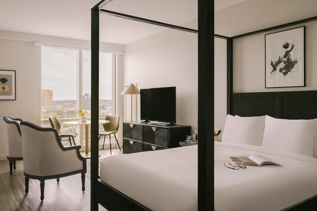 A hotel room with a bed, a television, a desk with a lamp, a chair, and a city view.