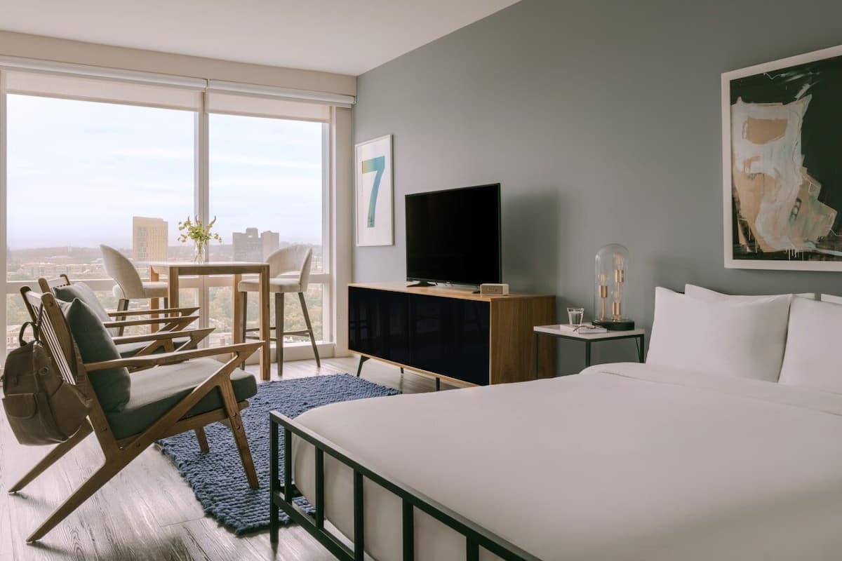 A hotel room with a bed, a television, a desk with a chair, and a large window offering a city view.