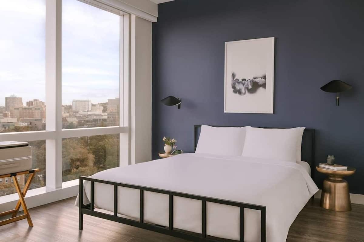 A bedroom with a bed, a city view, a framed artwork, and two wall-mounted lamps.