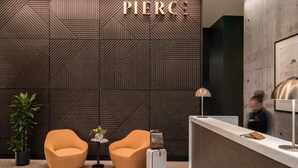 Un área de recepción con una pared de diseño geométrico, dos sillones naranjas, una pequeña mesa con un jarrón y un mostrador de recepción con una lámpara.