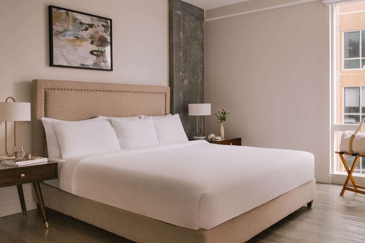 Ett hotellrum med en stor säng, sängbord, ett litet bord, en lampa, en vas med blommor och ett fönster med utsikt.
