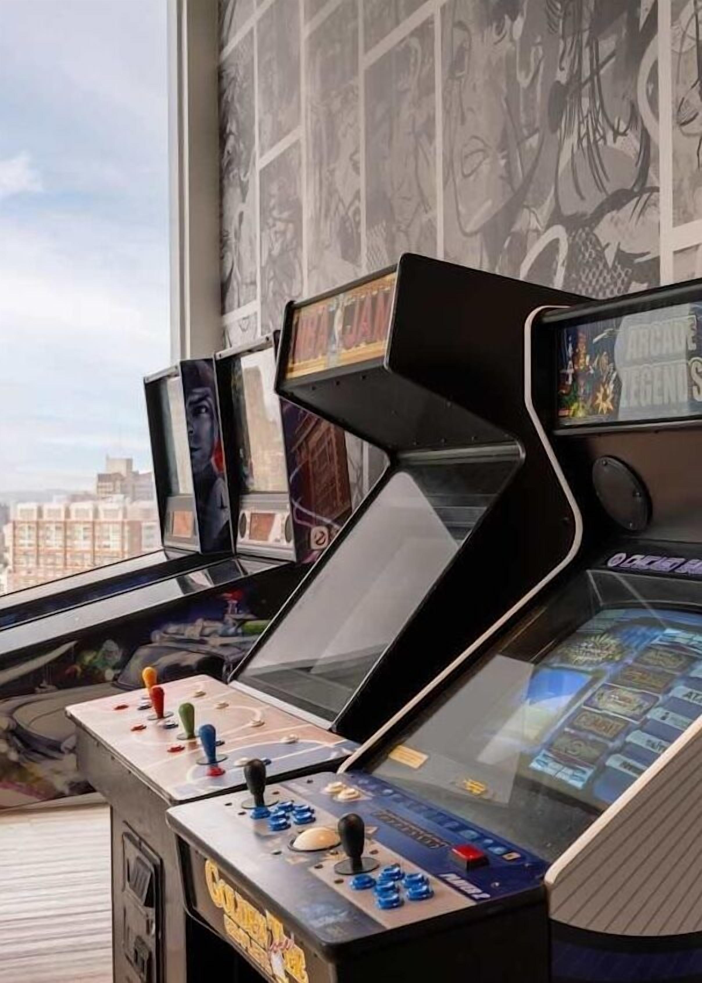 Des bornes d’arcade équipées de joysticks et d’écrans, placées près d’une fenêtre donnant sur la ville.