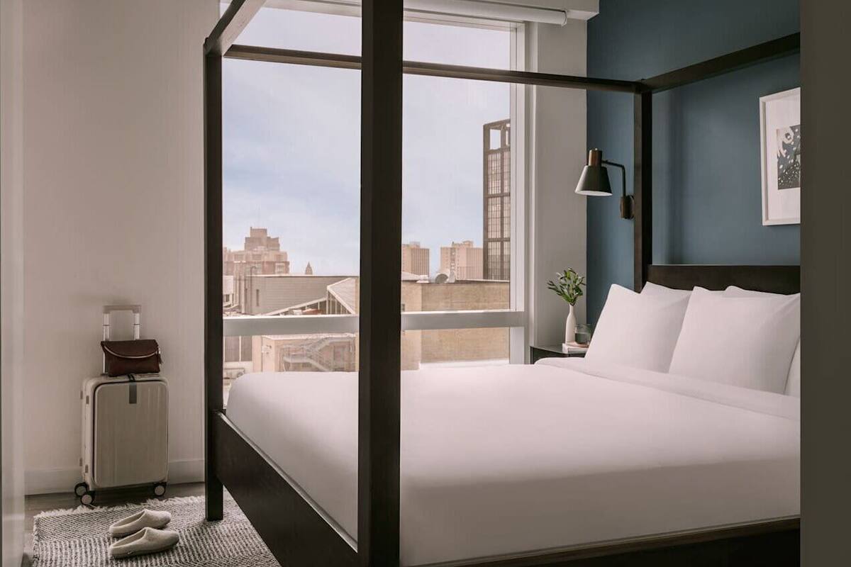 A hotel room with a large window, a bed, a nightstand, a lamp, and a suitcase.