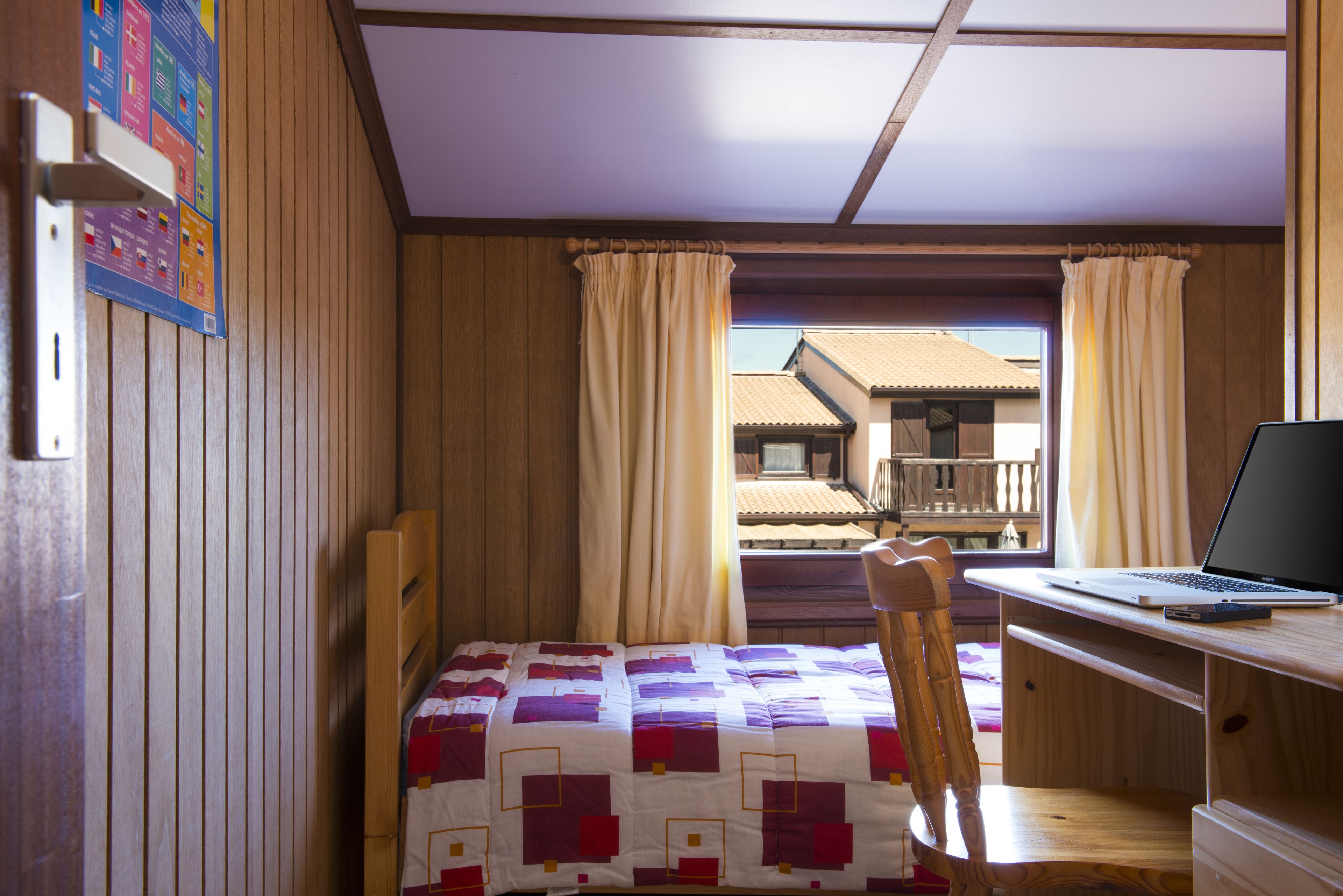A bedroom with a bed, a desk with a laptop, a chair, and a window with curtains.