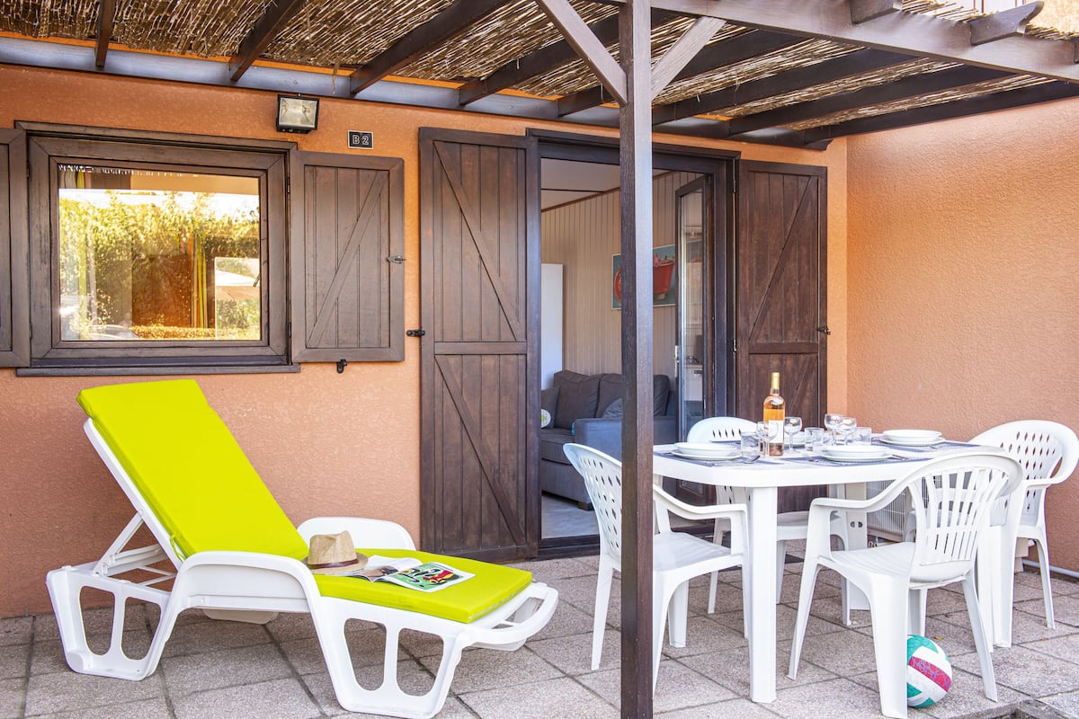 Un espace patio avec une table dressée pour les repas, un fauteuil de relaxation et une vue sur l’intérieur.