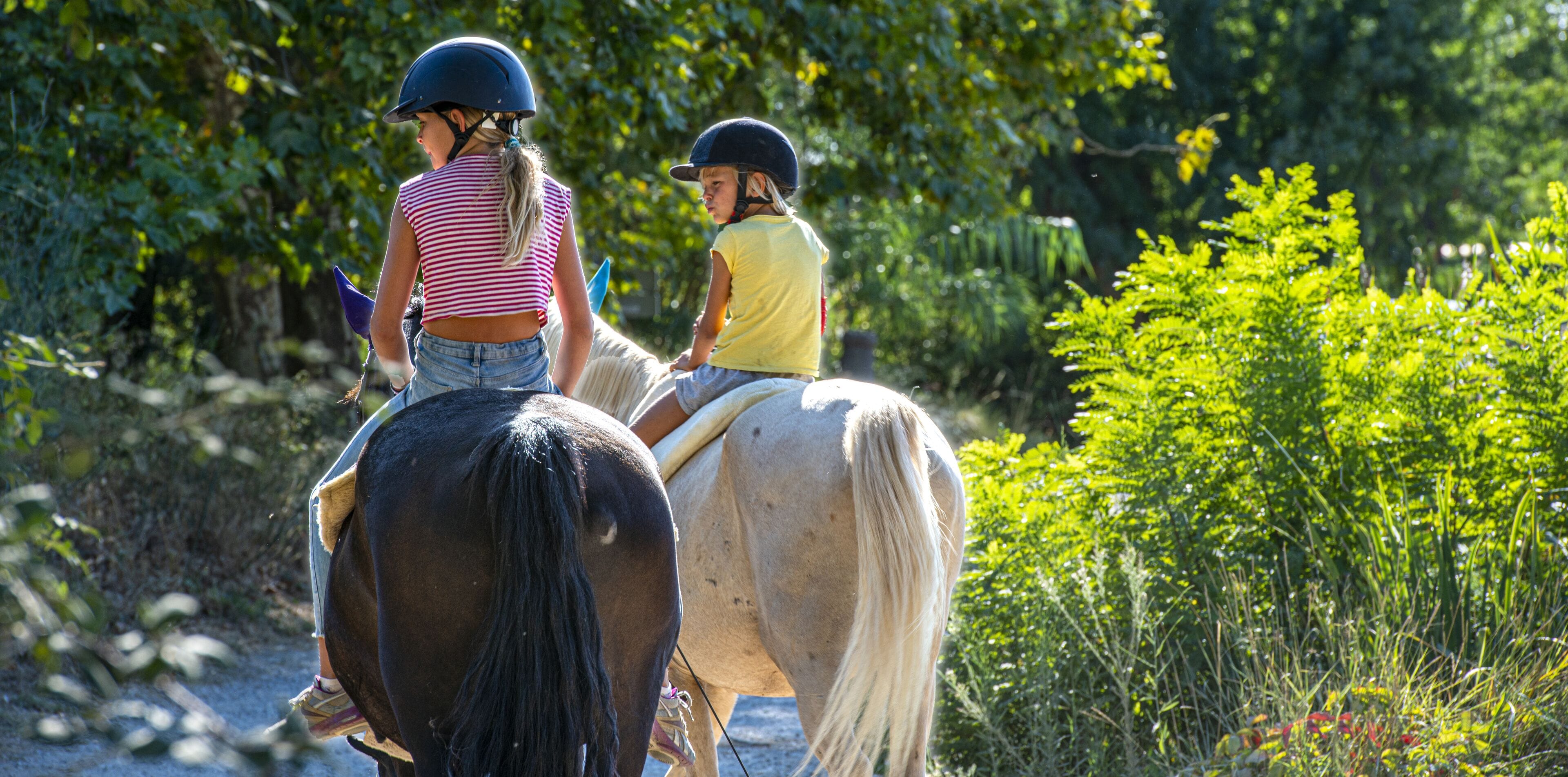 Dos niños montando caballos en un sendero de tierra rodeado de vegetación.