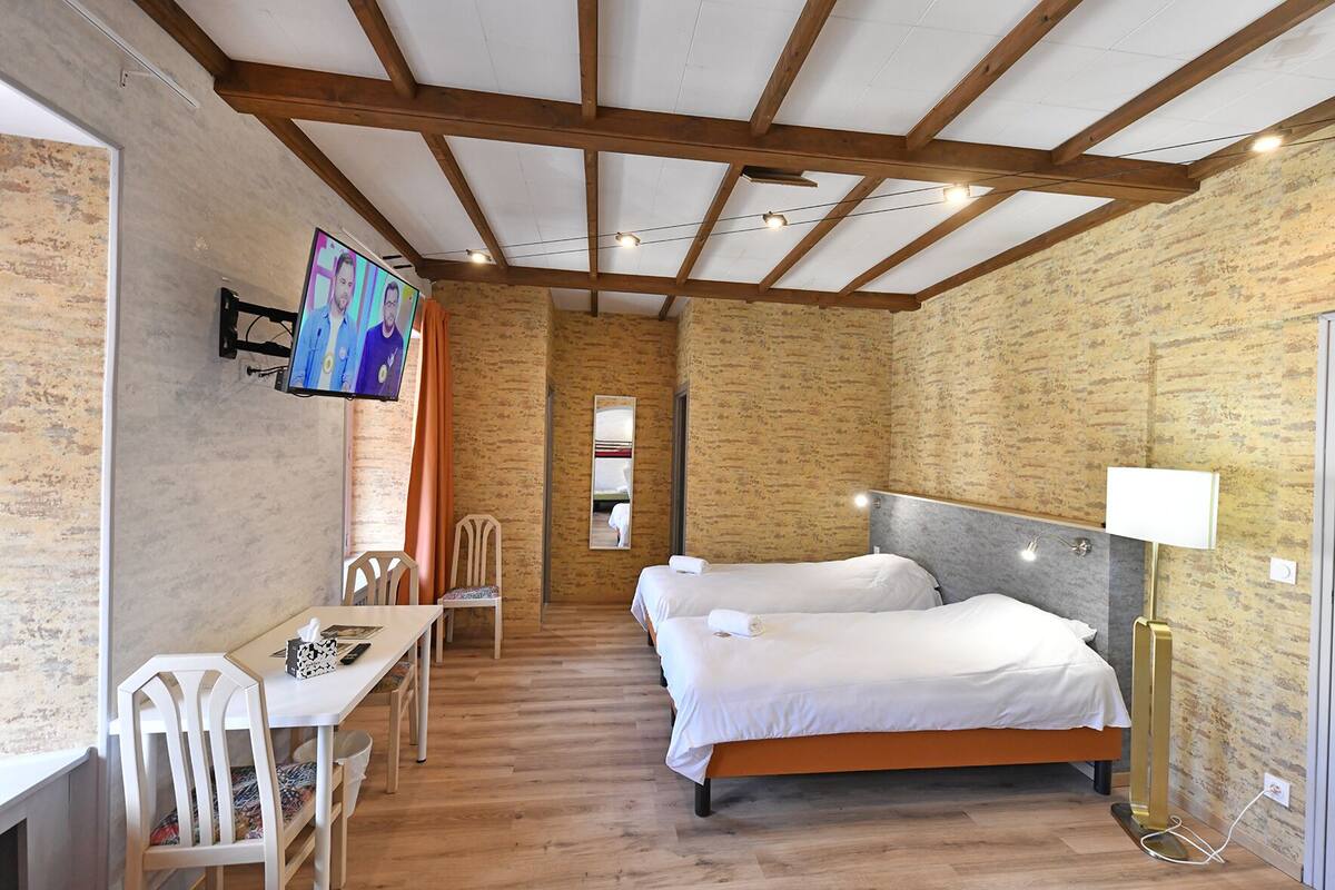 Une pièce avec un plafond en bois, deux lits avec du linge de lit blanc, un bureau avec des chaises, un téléviseur fixé au mur et un miroir.