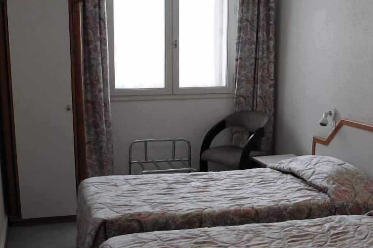 Une chambre avec deux lits, une chaise, un téléviseur posé sur un meuble et une fenêtre avec des rideaux.