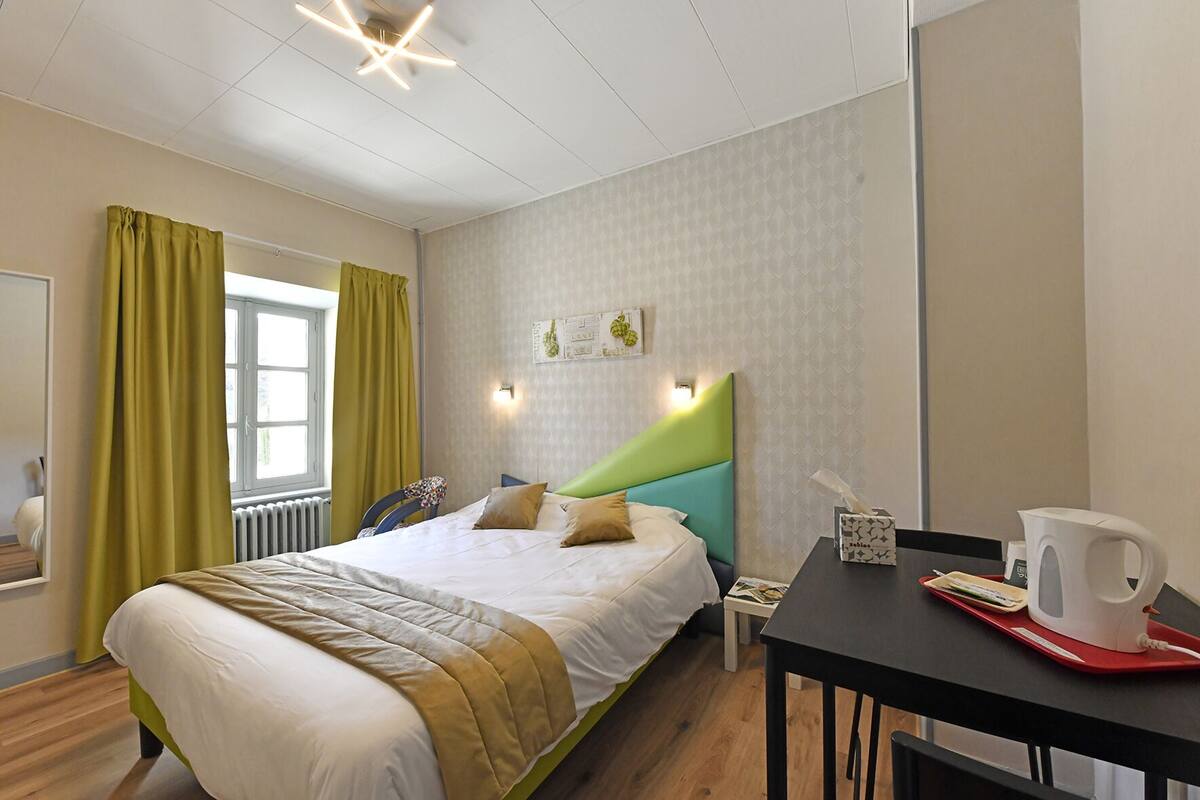 Une chambre d’hôtel bien rangée, avec un lit, une table sur laquelle se trouve une bouilloire, et une fenêtre avec des rideaux.