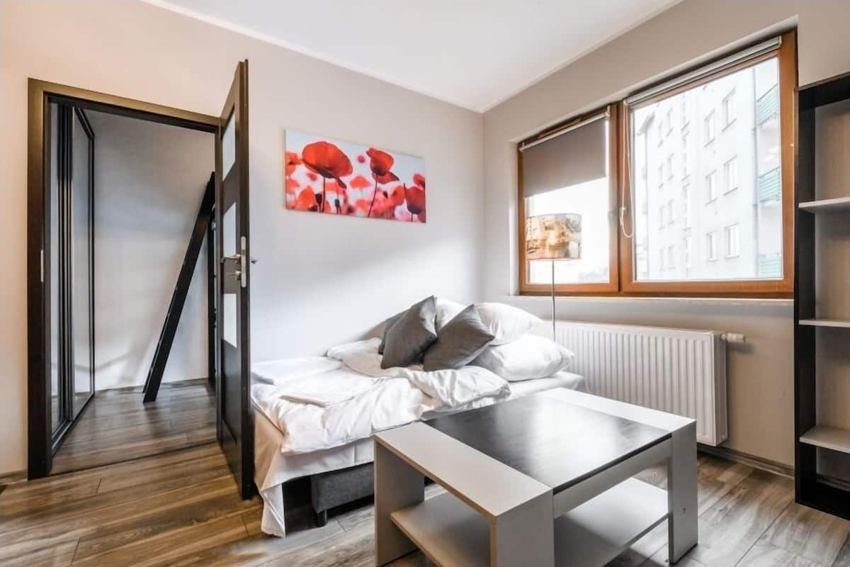 A bedroom with a bed, a table, a radiator, a window with a view of buildings, and a door leading to another room.