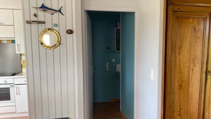 Una cocina con armarios blancos, encimera negra y suelo de baldosas. Se ve una puerta que da a un baño con paredes azules.