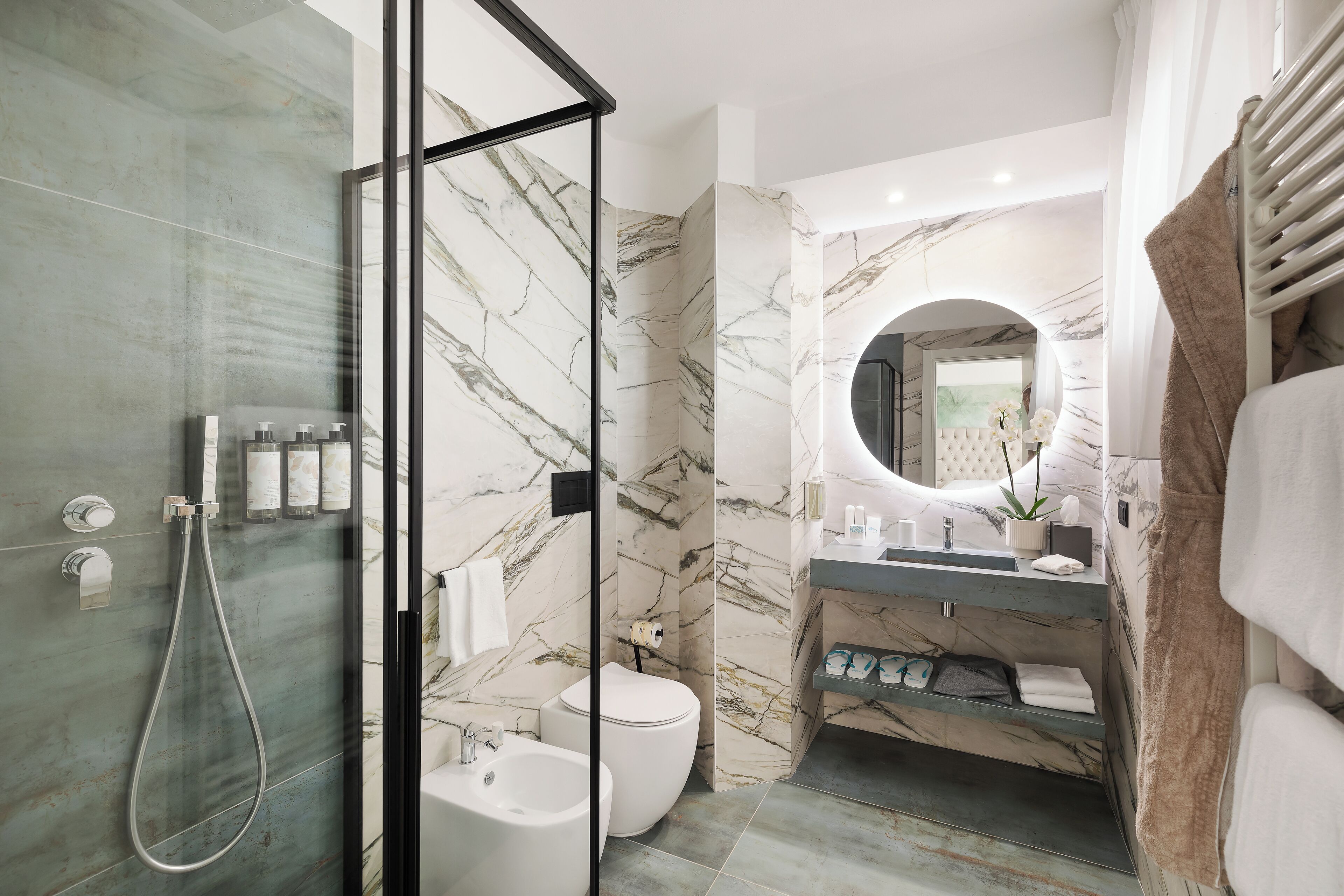 A modern bathroom with a glass-enclosed shower, a marble-tiled wall, a round mirror, and a built-in sink with a towel rack.