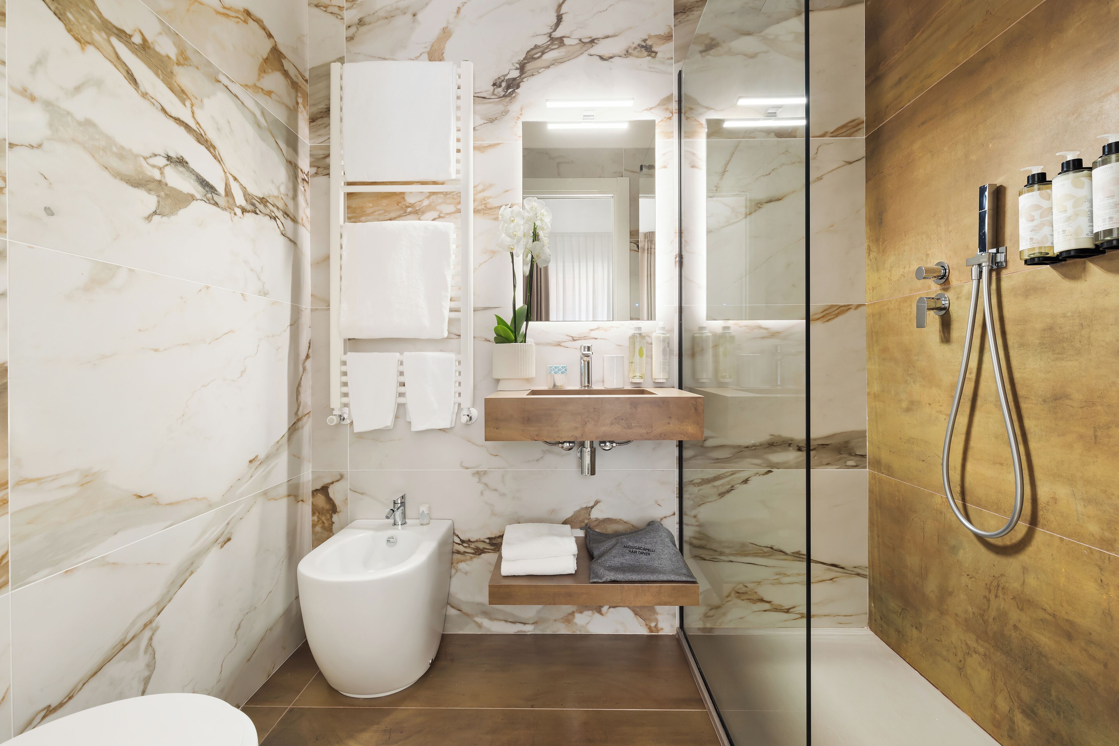 Un baño moderno con paredes de mármol, un bidé, un lavamanos con repisa de madera y una zona de ducha.