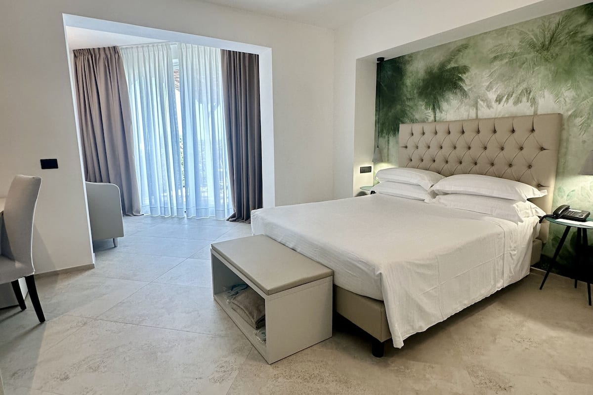 Ein modernes Hotelzimmer mit einem großen Bett, einer Sitzecke mit einem Sessel und einem kleinen Tisch sowie einem Ausblick durch ein großes, mit Vorhängen versehenes Fenster.
