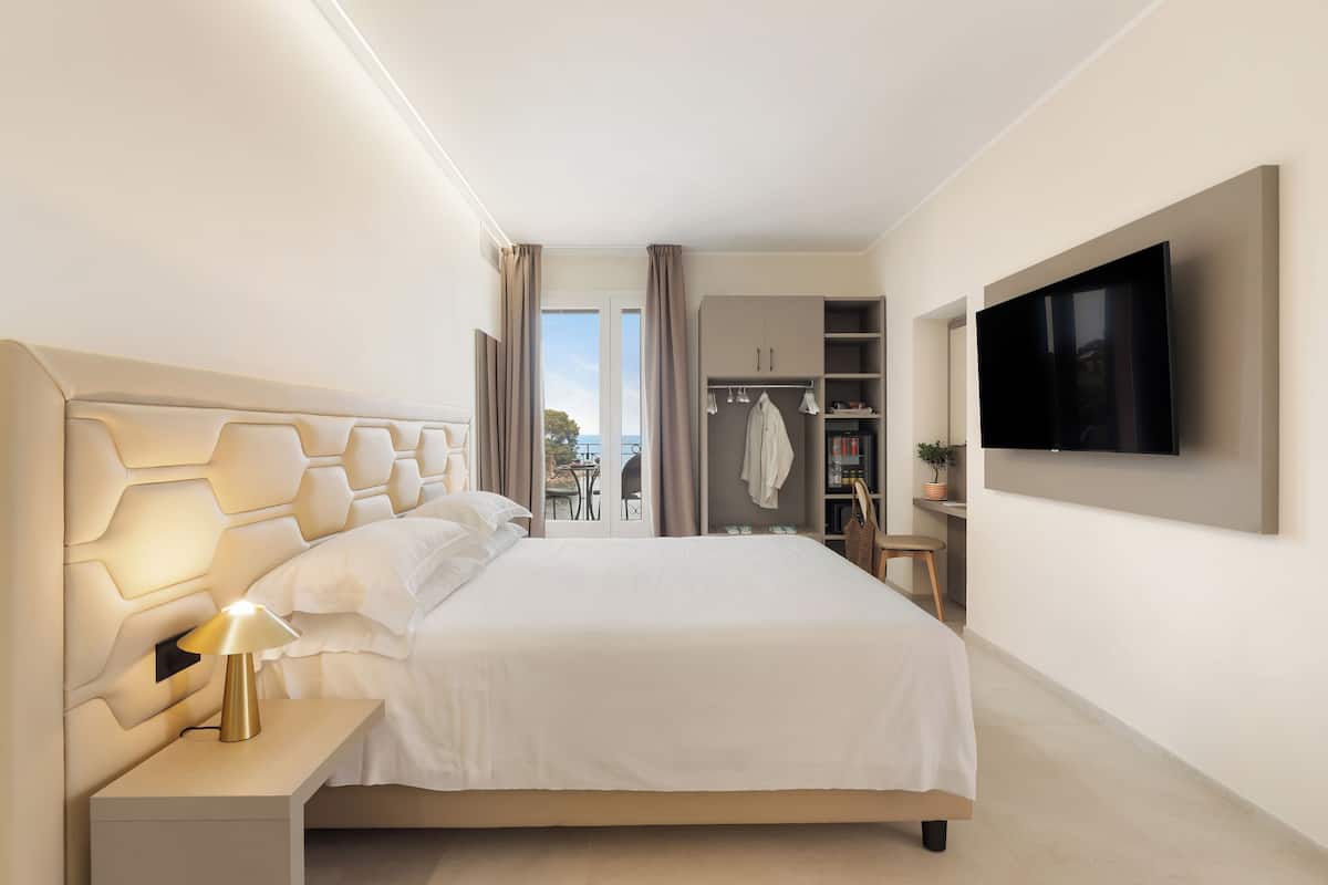 Ein modernes Hotelzimmer mit einem großen Bett, einem Nachttisch mit Lampe, einem Kleiderschrank, einem an der Wand befestigten Fernseher und Blick ins Freie durch das Fenster.