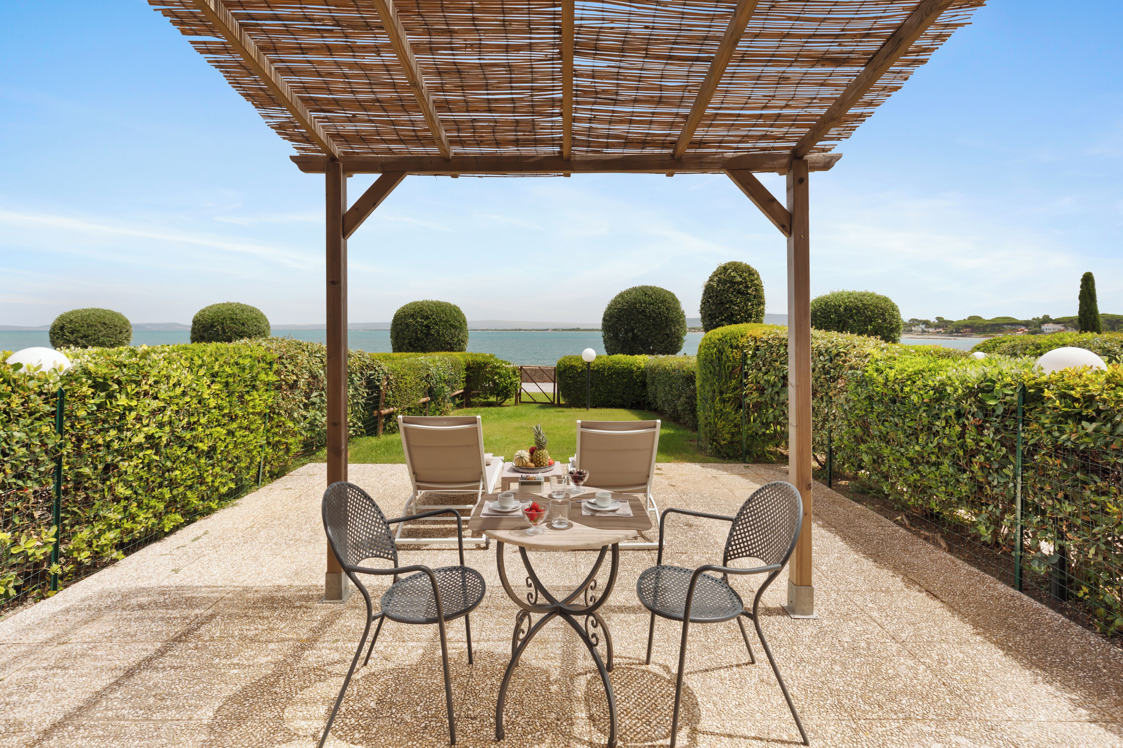 A covered outdoor dining area with a table and chairs, overlooking a scenic body of water and neatly trimmed hedges.