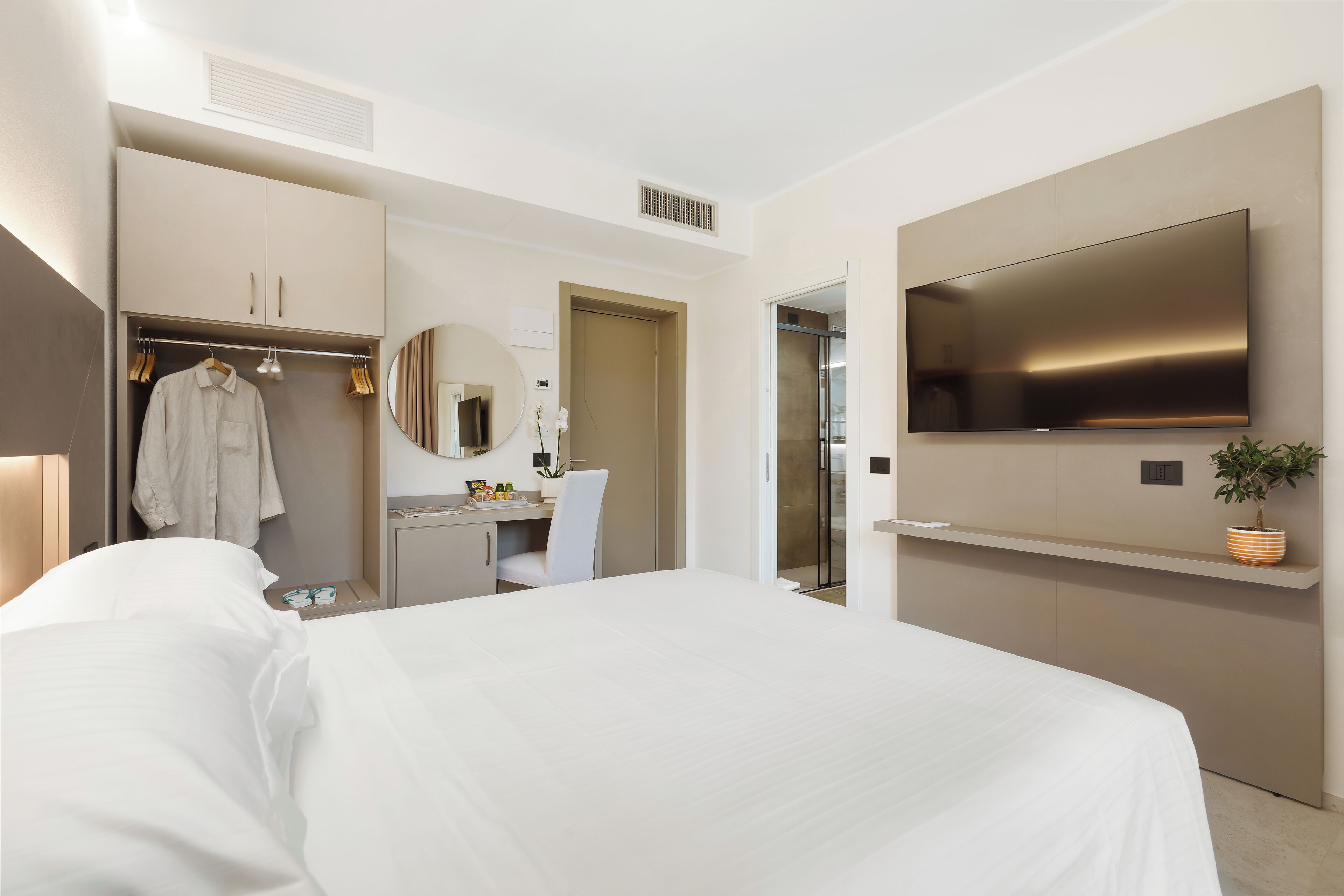 A modern hotel room with a bed, a desk, a wardrobe, a mirror, a television, and a potted plant.