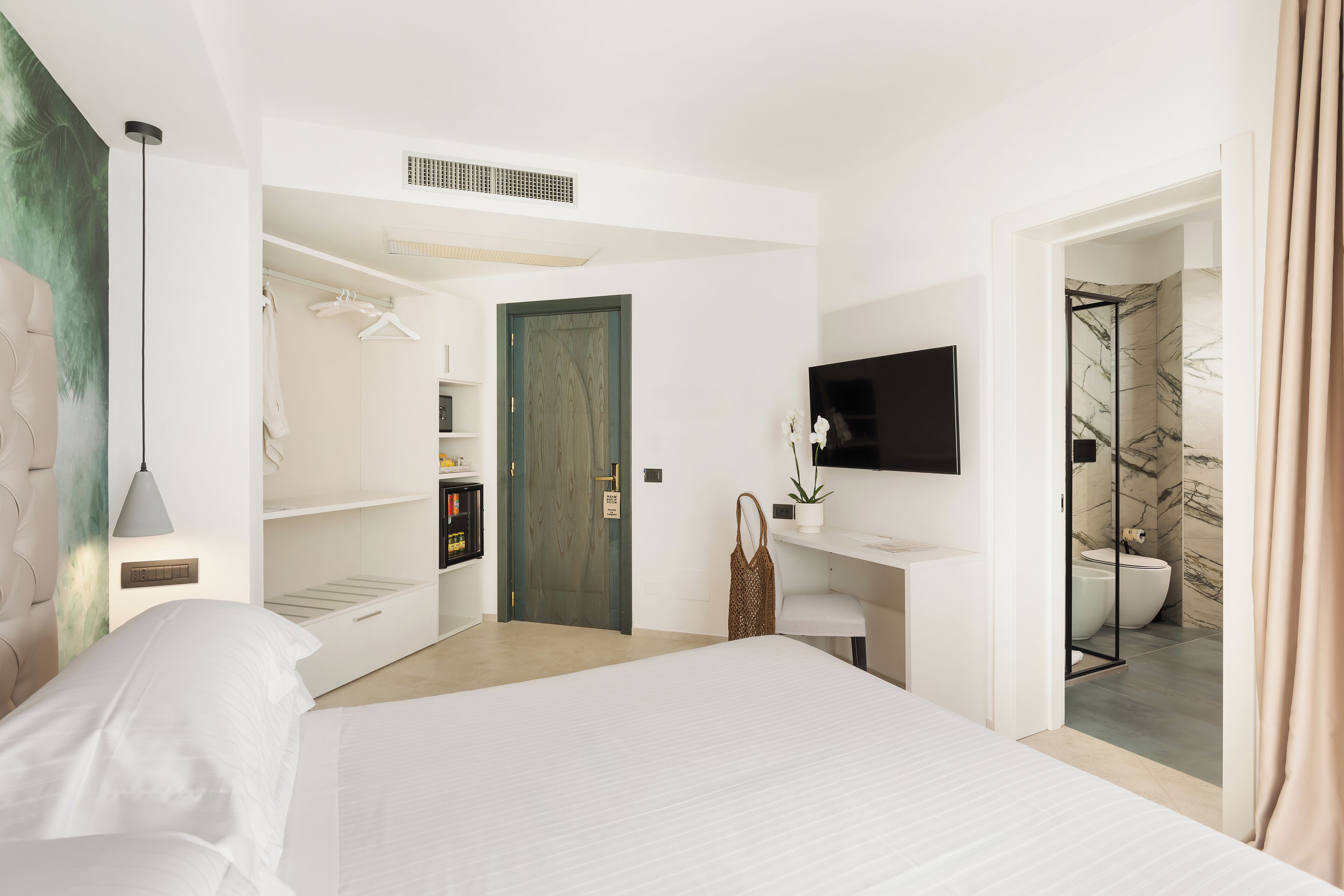 A modern hotel room with a bed, a TV, a desk, a wardrobe, and a bathroom with a marble shower.