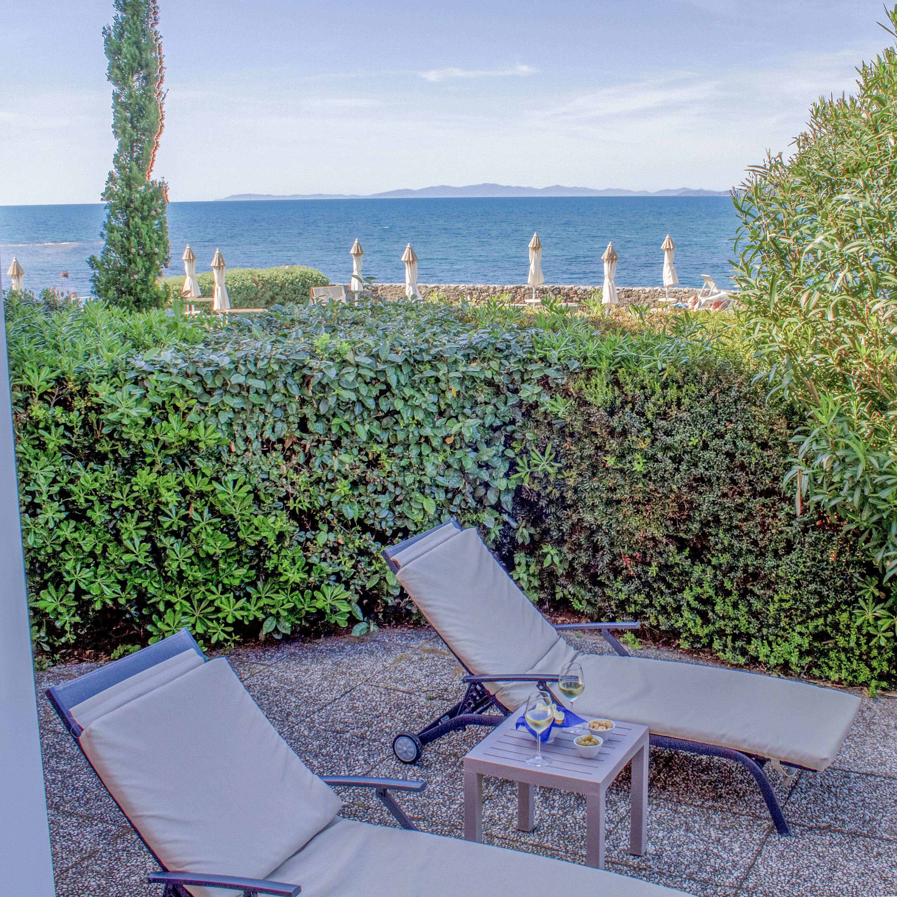 A terrace with two lounge chairs, a small table, and a view of the sea and mountains.