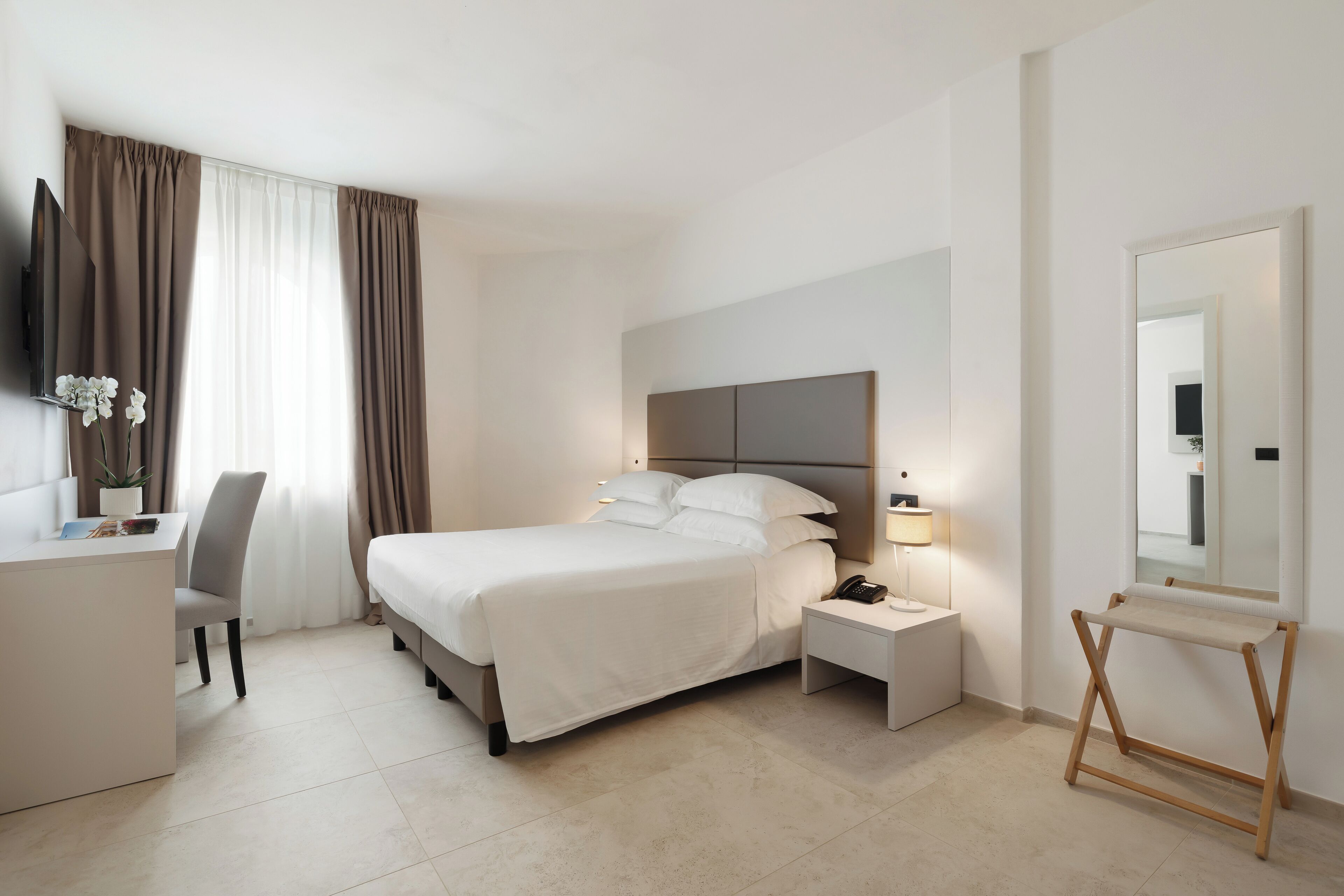 Una camera d'albergo moderna con un letto, un comodino, una lampada, una scrivania, una sedia e un grande specchio.