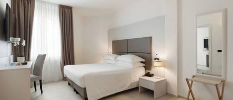 A modern hotel room with a bed, bedside table, lamp, desk, chair, and a large mirror.