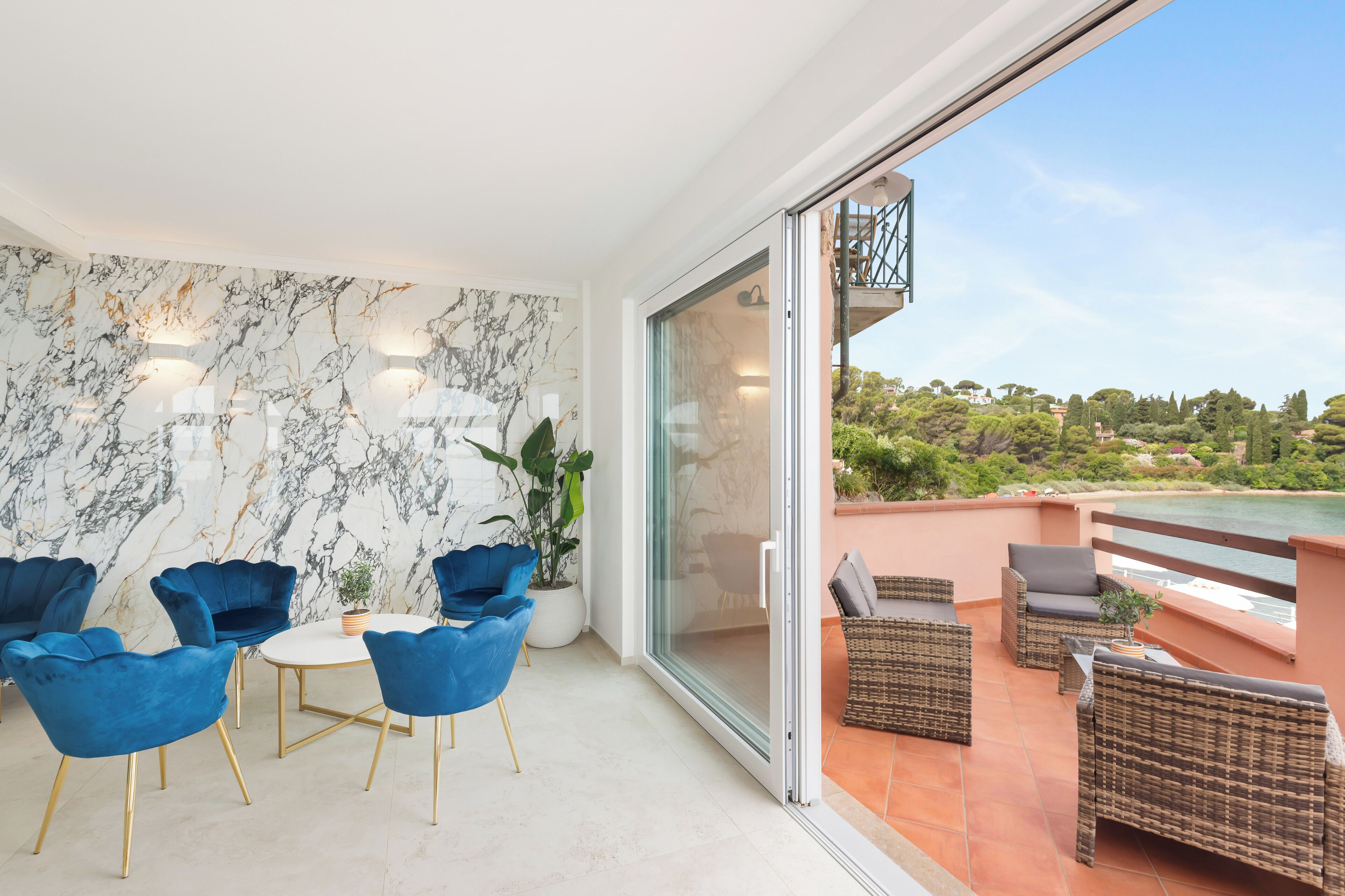 Een modern interieur met een marmeren wand, blauwe stoelen, een ronde tafel en een balkon met rieten meubels met uitzicht op een prachtig landschap.