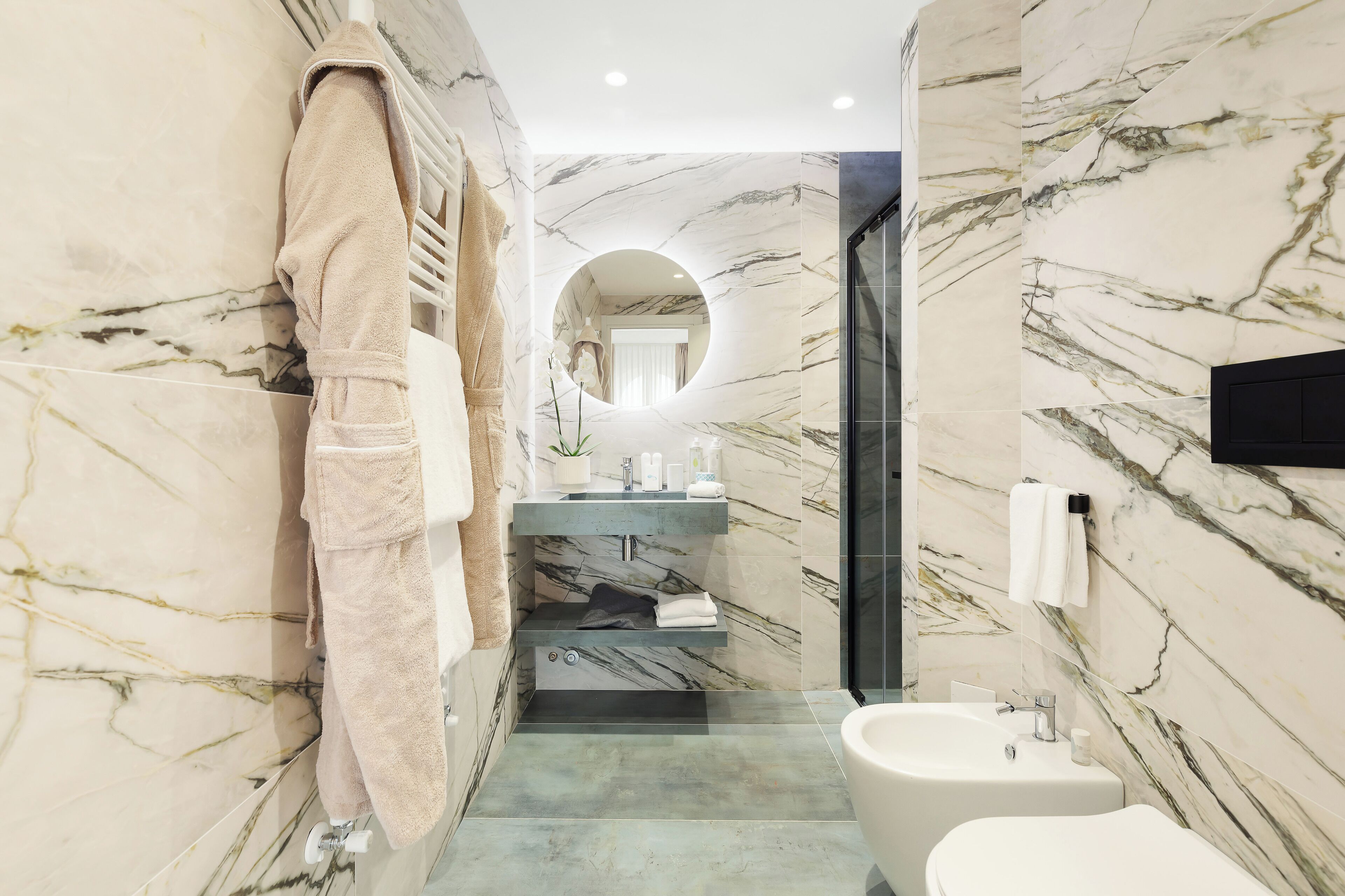 A modern bathroom with marble walls, a round mirror, a sink, and a bidet.