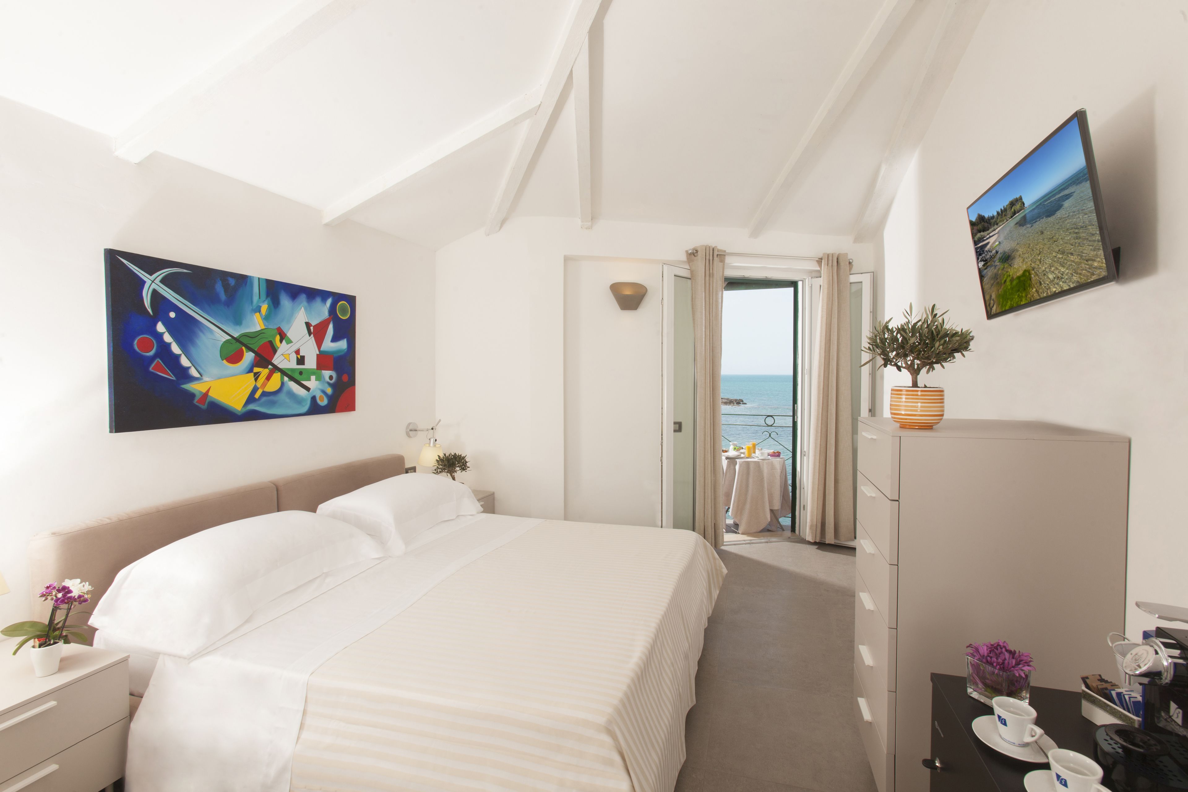 A hotel room with a large bed, a painting, a potted plant, and a view of the sea through an open door.