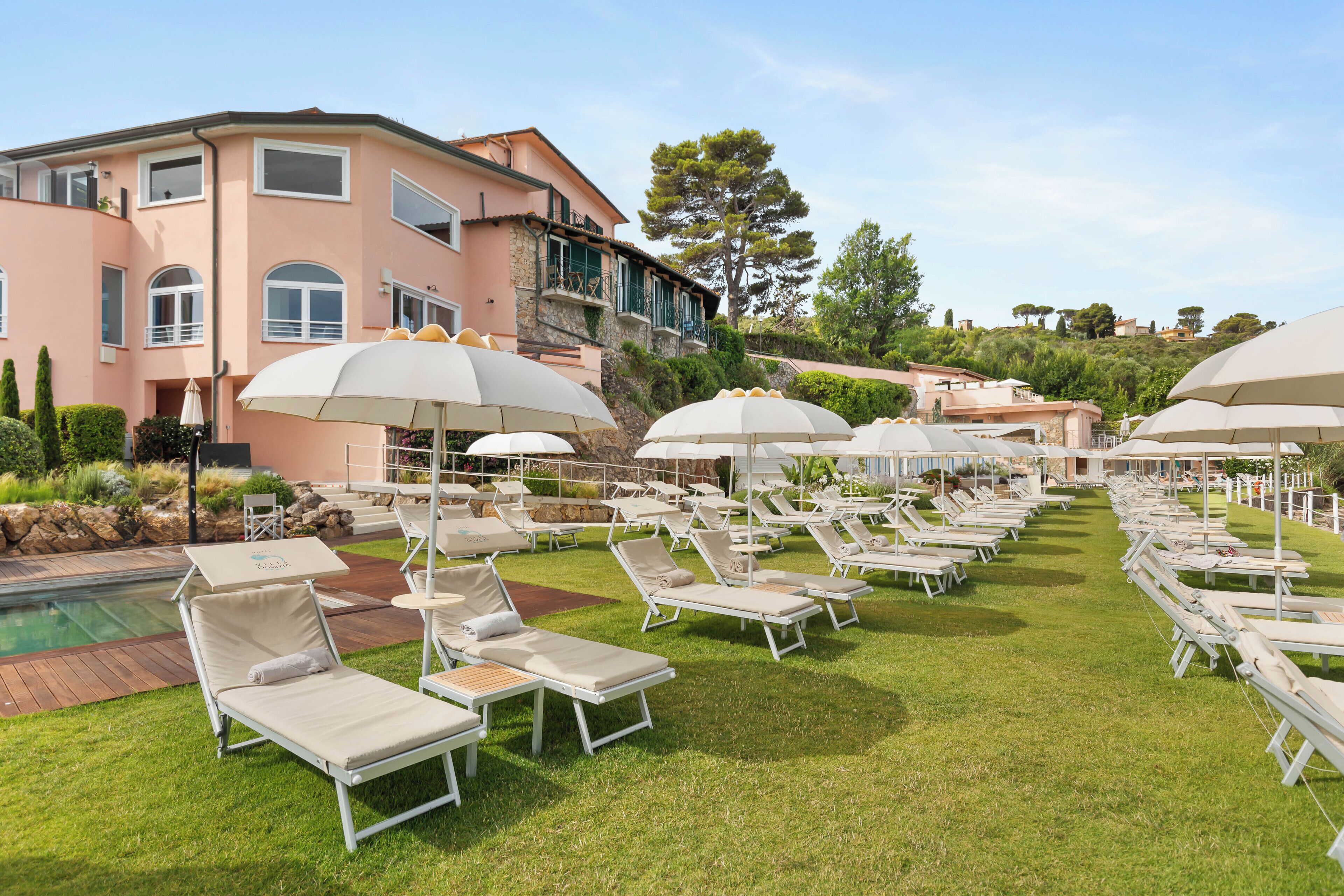 A poolside area with multiple sun loungers and umbrellas, a pool, and a large building in the background.