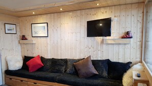 Um quarto aconchegante com parede de madeira, um sofá preto com vários almofadas, uma televisão montada e uma pequena mesa de madeira.