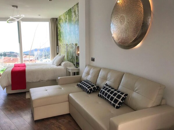 A modern hotel room with a bed, a beige sofa, and a large window offering a city view.