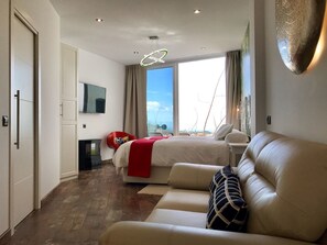 A modern hotel room with a large bed, a sofa, a TV, and a view of the sea.