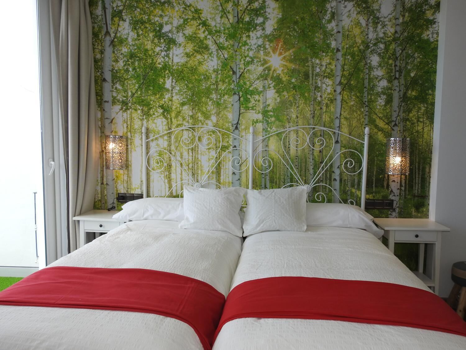 A double bed with white and red bedding, a metal headboard, and a wall mural of trees.