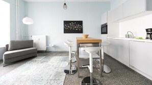A modern kitchen with white cabinets, a wooden table, and bar stools. There is a radiator, a floor lamp, and a black artwork on the wall. - Primeflats - Apartment Weissensee (Berlin)