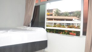 Quarto com cama, janela e vista para um edifício com varandas e plantas.