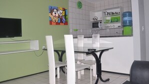 Uma sala de jantar com mesa de vidro e cadeiras brancas, uma TV fixada na parede e uma área de cozinha com micro-ondas e bebedouro.