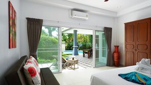 A hotel room with a bed, a sofa, a sliding glass door leading to a patio with a pool, and a wooden wardrobe.
