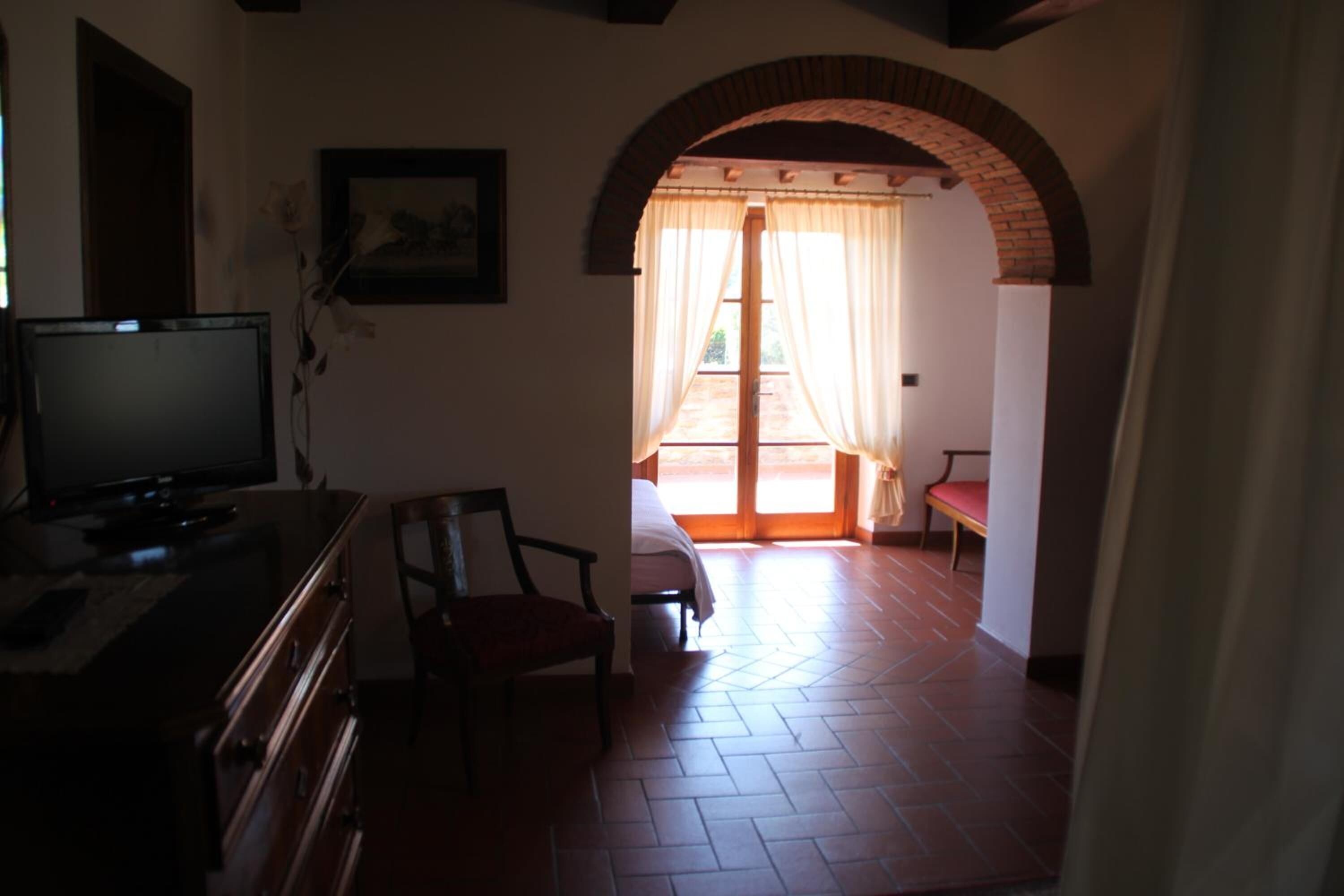 A room with a television on a dresser, a chair, a bed, and a view through an arched doorway to an outdoor area.