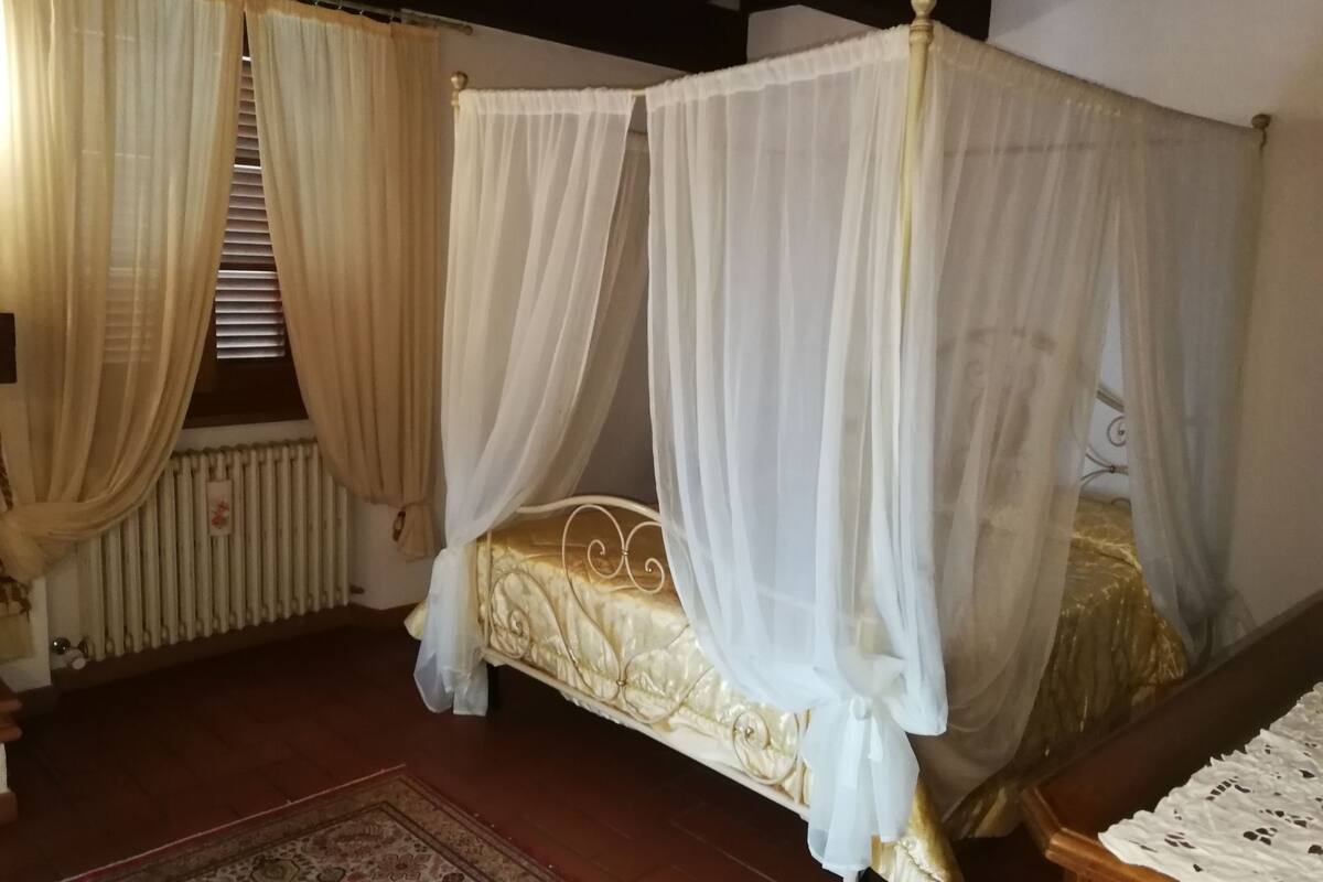 A four-poster canopy bed with a gold headboard, a wooden floor, a patterned rug, and a white lace tablecloth.